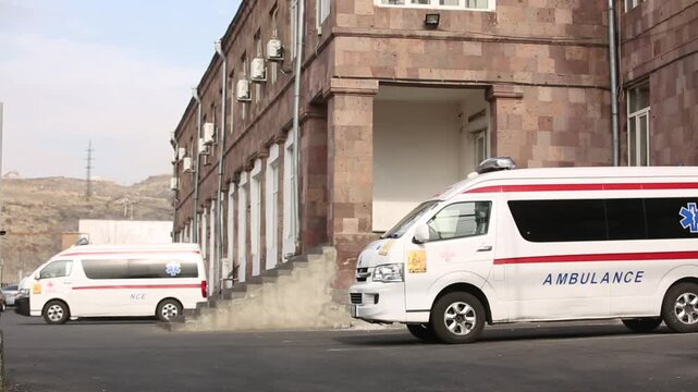 A white ambulance is parked outside a hospital building. In the background, a person walks up stairs. The emergency medical vehicle stands ready to respond to urgent calls.