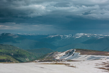 Dramatic landscape with small lake among snowfields, thawed ground, forest and snow cornice on long spur with view to big snowy mountain range far away under cloudy sky. Sunlight and shadows of clouds © Daniil