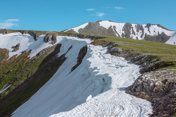 Scenic alpine sunlit view along green grassy ridge with white snow cornice and rocky sheer crags in bright sun under blue sky. Awesome landscape with sharp rocks and snowy mountain range in sunny day. © Daniil