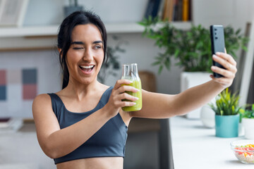 Fitness woman drinking a healthy green juice while taking a selfie with smartphone in the kitchen at home