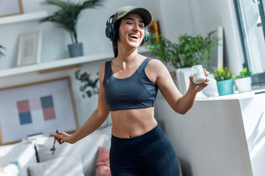 Sporty woman eating yogurt while dancing and listening to music with headphones at home