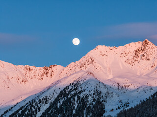 Full moon rises above jagged Dolomites in northern Italy at sunset. Pink alpenglow lights snow peaks, dark conifers and a lone chalet mark the valley under a crisp sky.