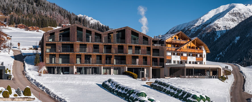 Modern alpine lodge in South Tyrol, Italy, with sawtooth roof and balconies sits in a snowy valley near Dolomite peaks, conifer forests, and a chalet as smoke drifts.