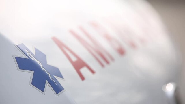 Close up shot with rack focus revealing the red word ambulance and blue star of life symbol printed on the side of a white emergency medical rescue vehicle.