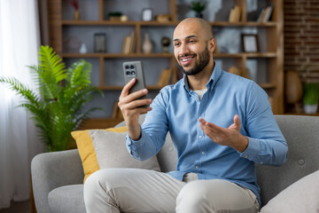 Smiling man sitting on a sofa in his living room, talking and actively gesticulating during a video...