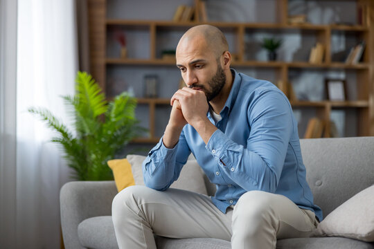 Upset young man sitting on sofa at home, looking pensive and worried, experiencing loneliness, stress and anxiety while reflecting on personal problems and needing support