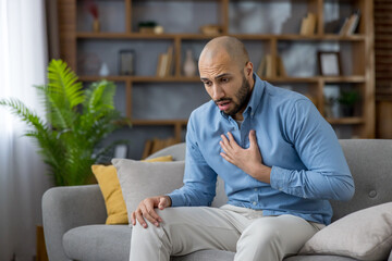 Man experiencing acute chest pain while sitting on a sofa in his living room, struggling with a...