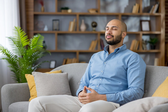 Young man relaxing on gray sofa at home, practicing mindfulness and deep breathing to find inner peace, calm, and balance during a quiet moment of self-care