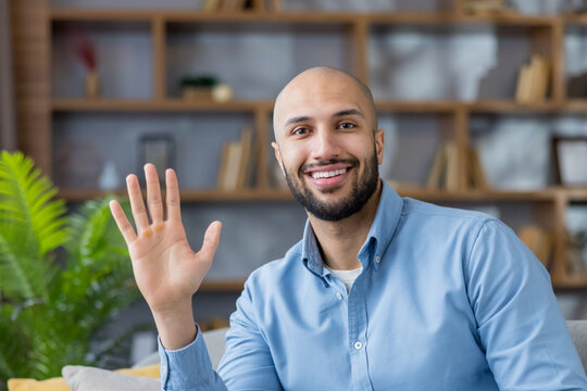 Young bald man with beard wearing a blue shirt, smiling at the camera and waving his hand in a friendly gesture, sitting indoors in a modern office or living room setting