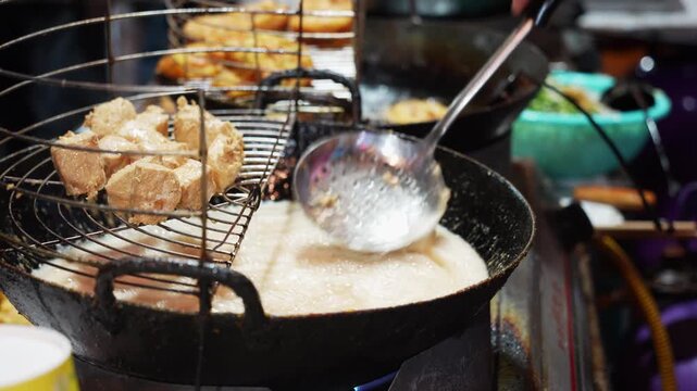 Street vendor frying tofu cubes in hot oil with a metal strainer and wok