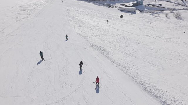 Aerial tracking shot following people skiing down a wide snow-covered slope at a winter mountain resort