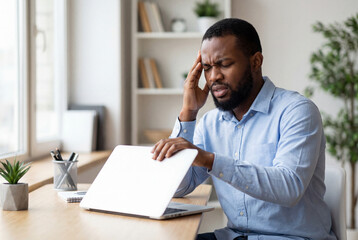 Obraz premium Stressed african american man touching temple and closing laptop lid at desk