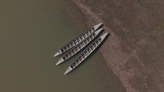Aerial top-down forward tracking shot of three empty traditional rowboats moored on a muddy lake shore