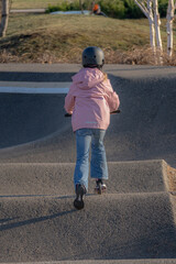 Fototapeta premium Young girl in a pink jacket and black helmet riding a scooter on a pump track at a skate park, back view, active outdoor lifestyle.