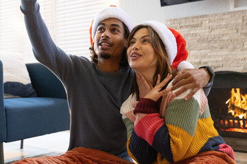Naklejka premium Couple sitting on blanket in front of fireplace wearing Santa hats and taking selfie