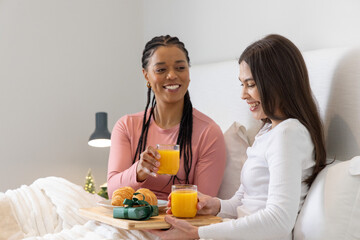Two women sitting on bed smiling, holding wooden breakfast tray with orange juice and croissants © wavebreak3