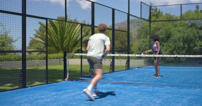 Diverse pair in sportswear playing on blue court with paddles for fun, man initiating forehand