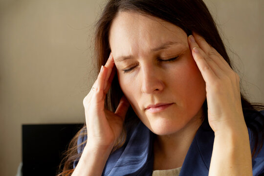 Woman with brown hair touching her temples indoors, headache or stress, closed eyes, blue shirt, home environment, daylight. health and discomfort, daily life.