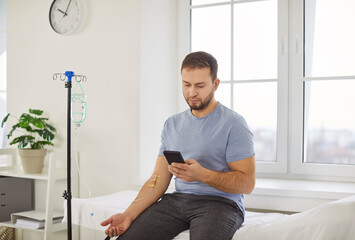 Man with IV drip infusion, patient sitting on hospital bed using smartphone while receiving...