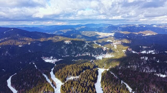 Tall trees cover the mountains and valleys with snow on the ground. The scenery shows winding paths through the forest. Clouds drift across the sky on a clear day.