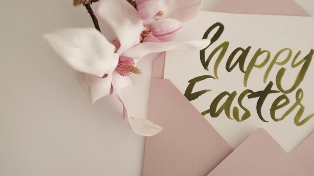 Happy Easter calligraphy lettering painted with green watercolor. Easter holiday greeting message in a pastel pink envelope with blooming magnolia flower. 