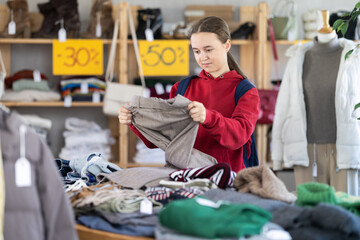 Focused young teen female client carefully choosing pants or stylish trousers in modern clothing...