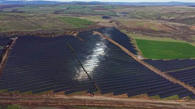 Wide view of a solar farm with many solar panels arranged in rows on open land. The scene shows green fields and hills in the distance under clear skies during the day.