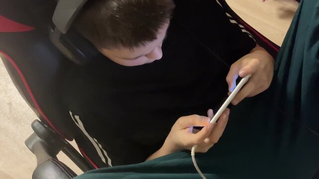 A 12-year-old boy sits in the evening wearing headphones, absorbed in his smartphone, the screen lighting his focused face as he listens and plays. Mobile games.