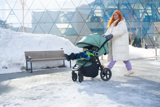Mature 43 year old red haired mother pushing mint baby stroller with 14 month old son while walking in city on sunny winter day.
