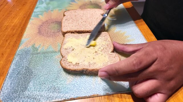 Male hands spreading butter on two slices of whole wheat bread with a knife on a glass cutting board. Preparing a simple sandwich