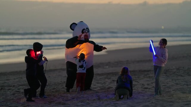 Children dancing with inflatable panda mascot on beach at sunset