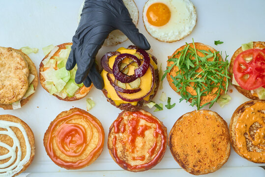 Chef's gloved hand assembling gourmet burgers, layering melted cheese, pickles and crispy red onions on grilled patties with fresh lettuce, tomato and sauces
