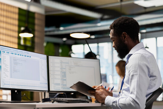 Programmer reviews documentation while coding on PC in modern office. Software developer cross referencing paperwork and source code at workstation to ensure accurate implementation