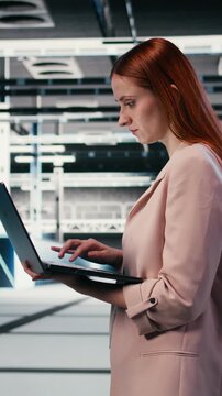 Vertical video Woman in server farm using laptop to improve electronics optimizations. Female technician walking in data center using notebook to monitor system performance, identifying bottlenecks