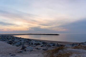 Obraz premium Deserted sandy beach with rocky breakwater curving into calm sea under soft pastel twilight sky at early winter dawn. Concept of coastal solitude, off-season travel, raw nature and peaceful emptiness.