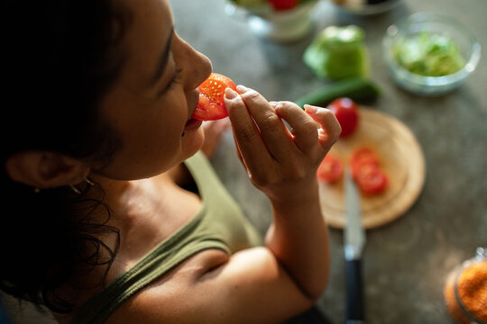 Young woman eating tomato slice in home kitchen