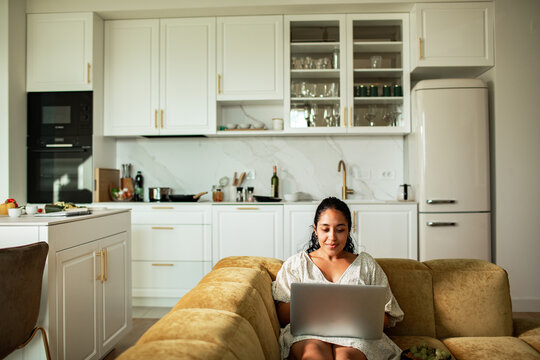 Woman using laptop on sofa in modern home kitchen