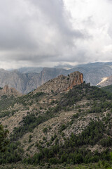 Fototapeta premium Moody clouds hover above the rugged Puig Campana region in Spain, highlighting rocky peaks and lush green forest, perfect for nature, travel, and adventure themes.