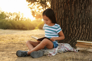 Naklejka premium Cute little girl reading book near tree in park