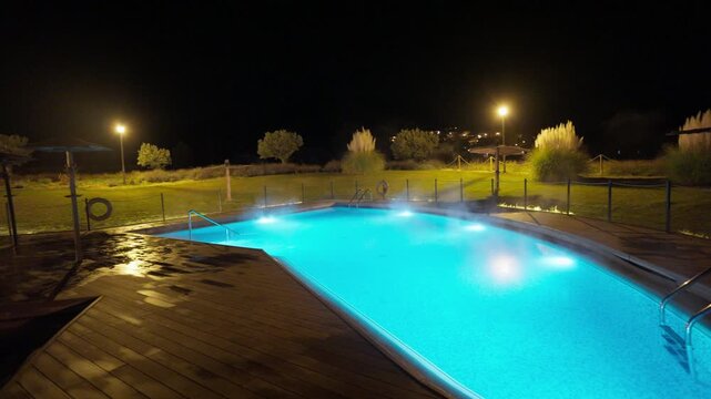A beautiful outdoor heated swimming pool glows vibrant blue at night, with steam rising from the warm water. The tranquil scene features a wooden deck, lush green grass, and distant ambient lights