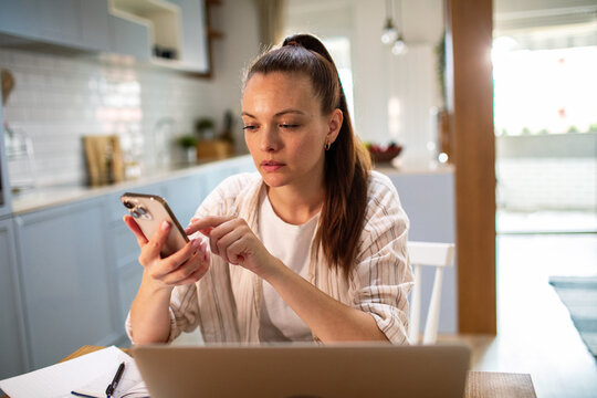 Young woman using smartphone at kitchen table while working from home