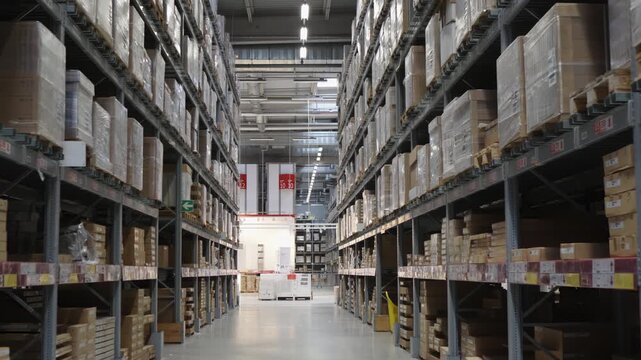Camera moves through a large, modern warehouse aisle. Tall metal racks are filled with cardboard boxes and inventory, showcasing logistics, storage, and supply chain management. No people.
