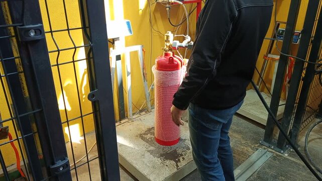 Technician performing safety inspection and weighing procedure of FM200 fire suppression cylinder in industrial facility following safety protocol.