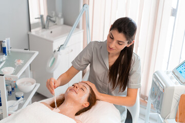 Cosmetologist in gray uniform applying facial mask to female client while lying on treatment bed in beauty salon. Professional skincare and cosmetology treatment concept