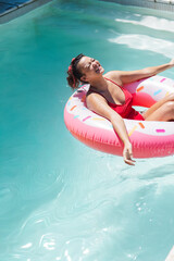 Woman 20s reclining on pink donut float in pool by coping, wearing red swimsuit, copy space