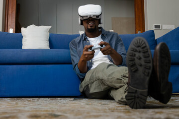 African American man sitting on floor leaning on blue sofa wearing white VR headset holding gamepad © wavebreak3