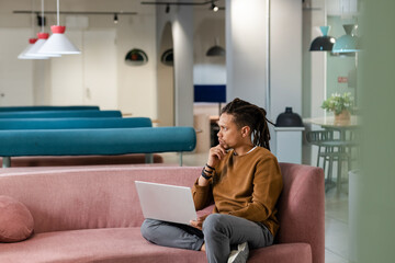 Obraz premium African American man sitting on pink sofa in co-working lounge holding silver laptop, copy space