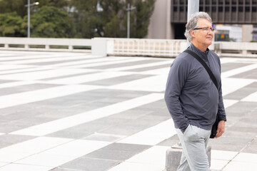 Mature man standing by pole and bollard on plaza wearing glasses carrying crossbody bag, copy space © wavebreak3