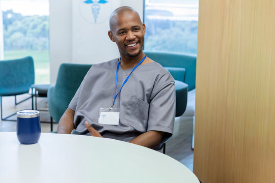 Man in gray scrubs sitting, smiling at staff lounge round table with tumbler and ID badge