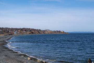 Topsail Beach, Newfoundland, has rolling waves, a shore lined with seaweed, and a rocky pebbled coast. The sunny day has a blue sky with clouds. There are houses and a church on the cliff.